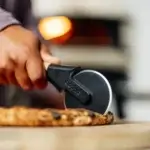 Mano usando el Gozney Pizza Cutter para cortar una pizza, con la rueda en primer plano y un horno encendido al fondo.