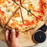 Pizza cortada en porciones sobre tabla de madera, con una mano levantando una porción y el Gozney Pizza Cutter al lado