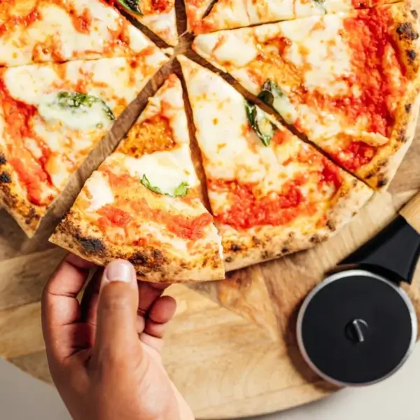Pizza cortada en porciones sobre tabla de madera, con una mano levantando una porción y el Gozney Pizza Cutter al lado