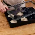 Manos levantando la tapa de una Gozney Dough Tray con bolas de masa sobre una mesa de madera.