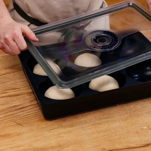 Manos levantando la tapa de una Gozney Dough Tray con bolas de masa sobre una mesa de madera.