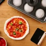 Pizza con tomate y albahaca junto a la Gozney Dough Tray con bolas de masa y una espátula sobre mesa de madera.