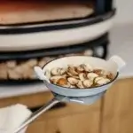 Pala Gozney Utility sosteniendo una fuente blanca con champiñones delante de un horno Gozney.