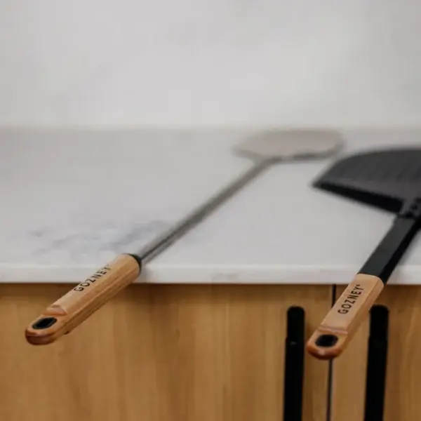 Pala Gozney Utility apoyada sobre una encimera junto a otro accesorio Gozney con mango de madera.