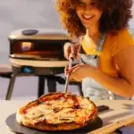 Persona de cabello rizado usando la pala Gozney Utility junto a una pizza y un horno Gozney.