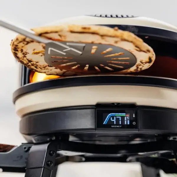 Pala perforada Gozney Balance levantando una pizza en un horno Gozney con llama y pantalla digital visibles.