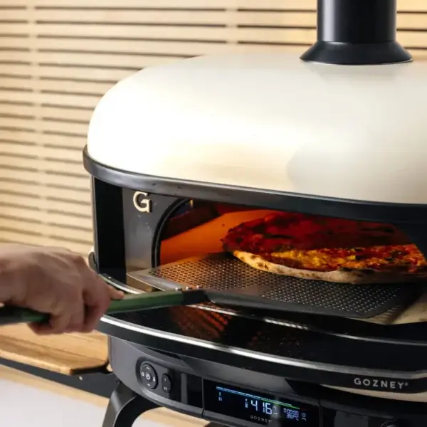 Mano sujetando una Gozney Pro Peel 16 XL para introducir una pizza en un horno Gozney encendido.