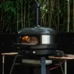Soporte Gozney Dome Gen 2 con horno Off-Black encendido en un jardín, accesorios en las alas laterales y vegetación de fondo.