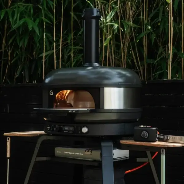Soporte Gozney Dome Gen 2 con horno Off-Black encendido en un jardín, accesorios en las alas laterales y vegetación de fondo.