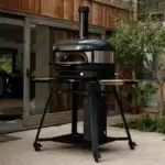 Horno Gozney Dome con arco napolitano sobre soporte con ruedas en una terraza exterior junto a madera y vegetación.
