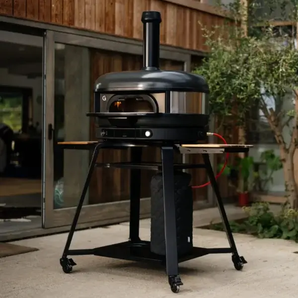 Horno Gozney Dome con arco napolitano sobre soporte con ruedas en una terraza exterior junto a madera y vegetación.