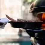 Close-up of a cast iron pan resting on the Gozney Dome Gen 2.0 XL front shelf, with a gloved hand cooking beside the oven.