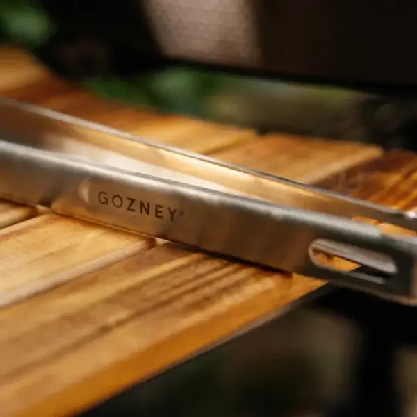 Detalle del logotipo Gozney grabado en las pinzas de fuego vivo sobre una tabla de madera.