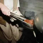 Manos usando las pinzas Gozney para sujetar carne sobre una parrilla.