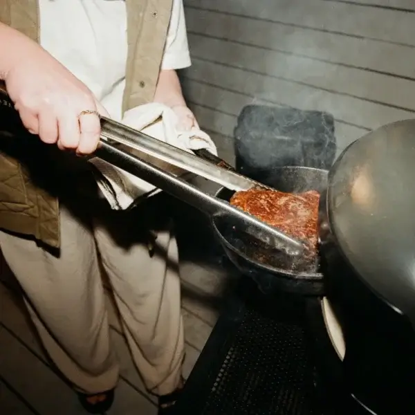 Manos usando las pinzas Gozney para sujetar carne sobre una parrilla.