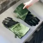 Par de guantes Gozney de ante verde y negro colocados sobre una mesa de acero, con una mano sujetando uno.