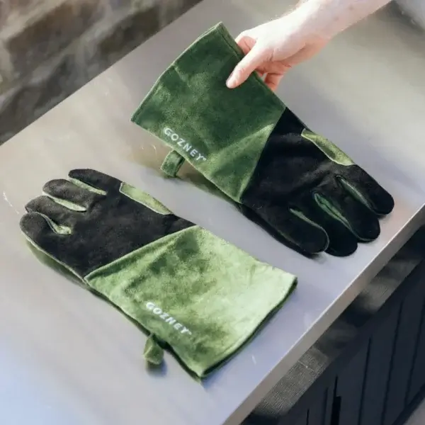 Par de guantes Gozney de ante verde y negro colocados sobre una mesa de acero, con una mano sujetando uno.