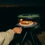 Mano usando la pala Gozney Venture para introducir una pizza en un horno Gozney al aire libre de noche.