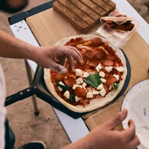 Manos preparando una pizza con pepperoni, queso y albahaca sobre la pala Gozney Venture.