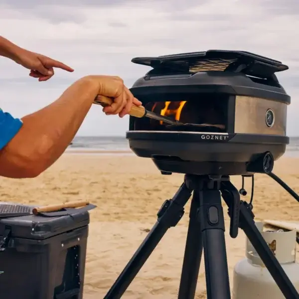 Manos usando la pala Gozney Venture en un horno portátil Gozney sobre trípode en un entorno de playa, con llama visible.