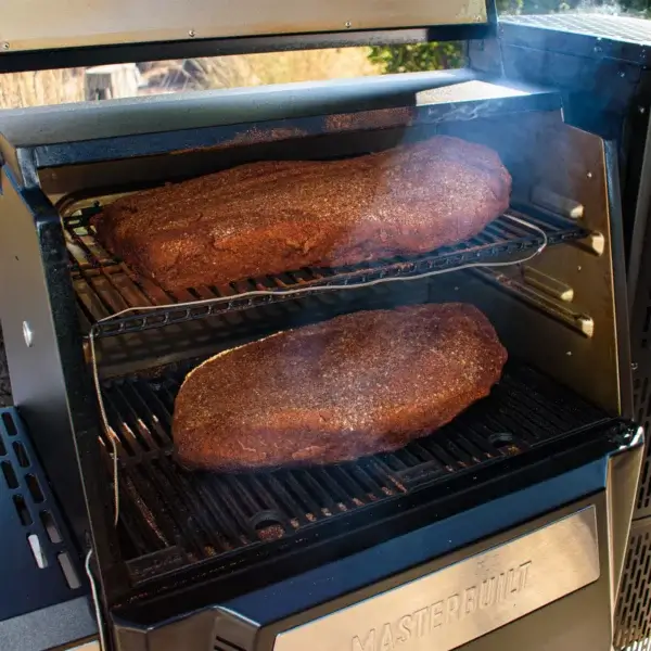 Dos piezas grandes de carne ahumándose sobre las rejillas de un equipo Masterbuilt Gravity Series con humo visible.