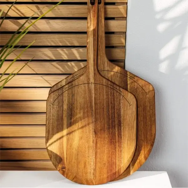 Pala Gozney de acacia de 12 pulgadas junto a otra pala de madera en un ambiente exterior con listones y luz natural.