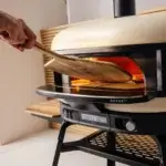 Una mano sostiene la pala Gozney de acacia de 16 pulgadas XL con una pizza mientras la introduce en un horno encendido.