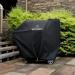 Masterbuilt Gravity Series 800 cubierta con funda negra en un patio exterior junto a vegetación y muro de piedra.