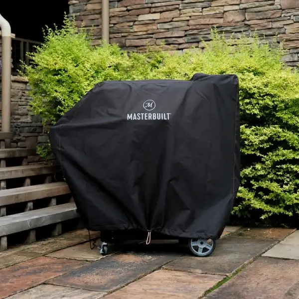 Masterbuilt Gravity Series 800 cubierta con funda negra en un patio exterior junto a vegetación y muro de piedra.