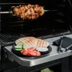 Carne cocinándose en un rostizador dentro de la parrilla, con pan, pepino, tomate y salsa servidos en la mesa lateral.