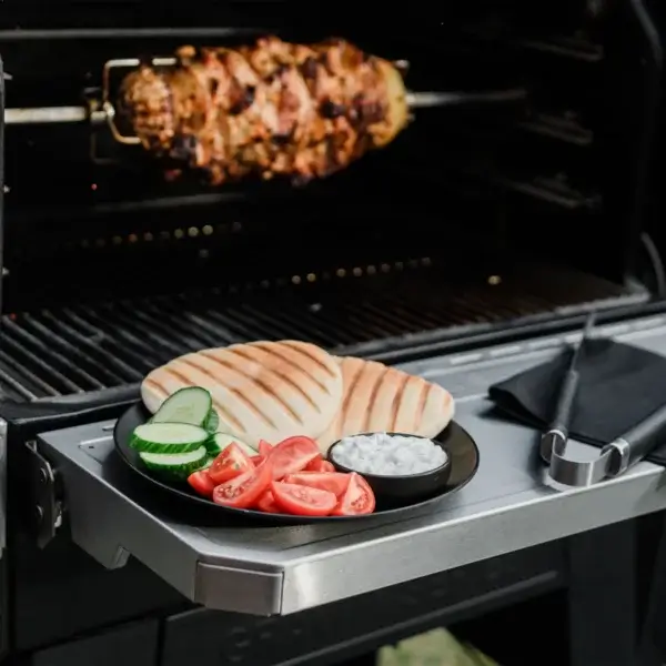 Carne cocinándose en un rostizador dentro de la parrilla, con pan, pepino, tomate y salsa servidos en la mesa lateral.