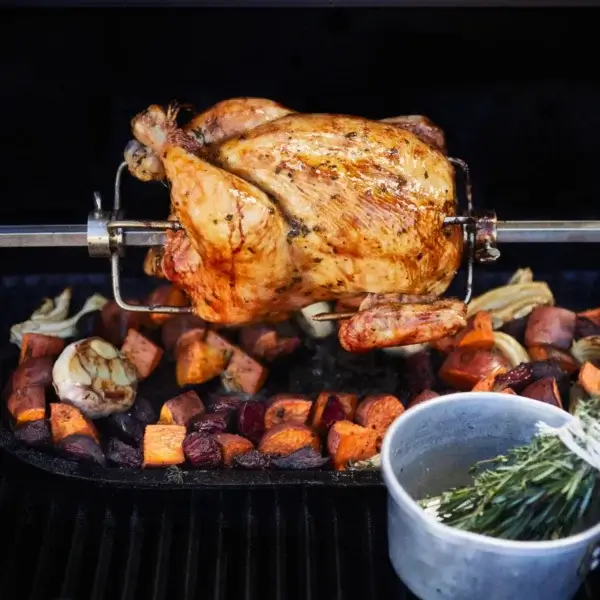 Pollo entero dorado girando en el rostizador sobre verduras asadas, con un cuenco de hierbas en primer plano.