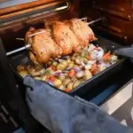 Pollos dorados girando en el rostizador sobre una bandeja con patatas, cebolla y otras verduras dentro de una parrilla.