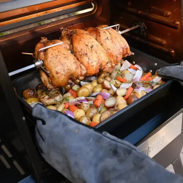 Pollos dorados girando en el rostizador sobre una bandeja con patatas, cebolla y otras verduras dentro de una parrilla.