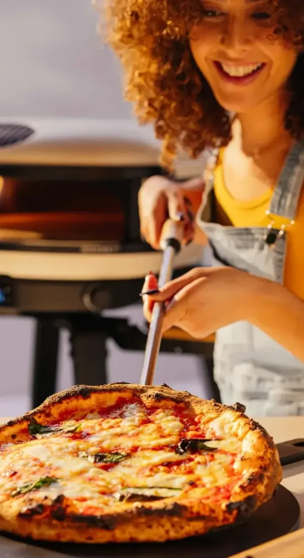 Persona de cabello rizado usando la pala Gozney Utility junto a una pizza y un horno Gozney.